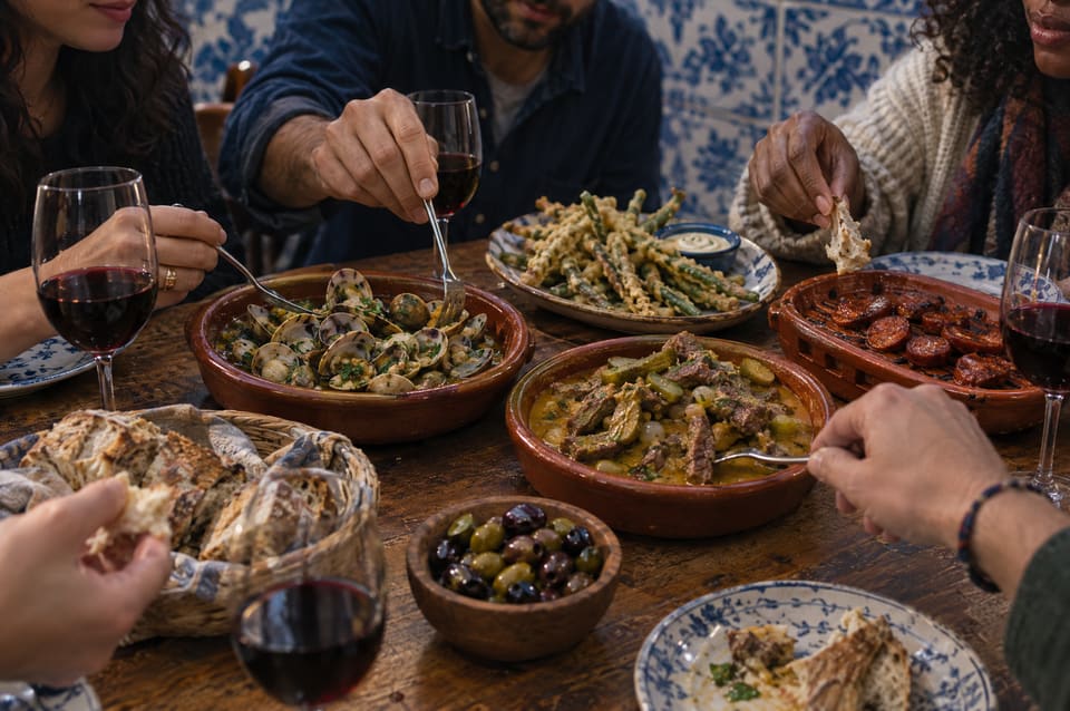 Friends sharing clams, beef pica-pau, tempura green beans, chorizo and olives on a wooden table with glasses of red wine.